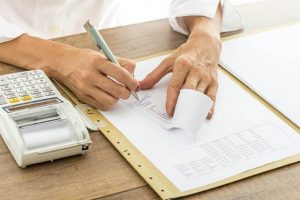 Foto de uma mão anotando em um bloco e com uma calculadora anotando os erros em notas fiscais