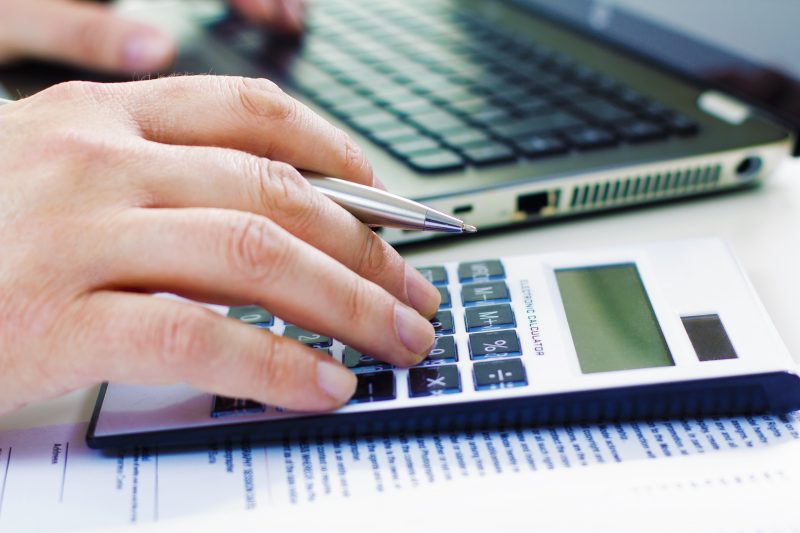 Foto de uma mão masculina digitando em um computador e fazendo conta de finanças pessoais em uma calculadora
