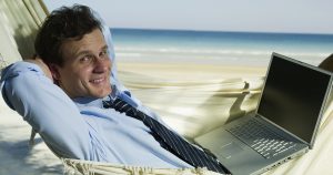 Foto de um empresário na praia com um notebook no colo, representando as férias de final de ano para empreendedores