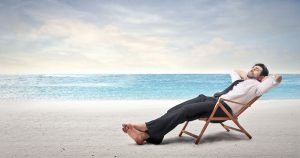 Foto de um homem vestido de camisa e calça social, sentado à praia, representando as férias coletivas