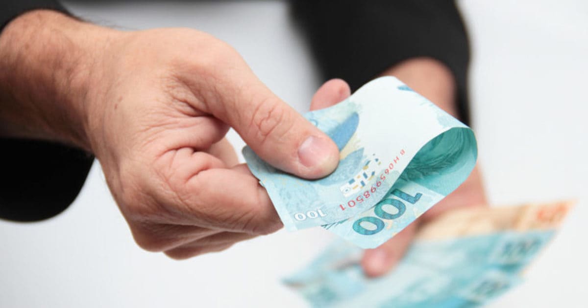 Foto de uma mão masculina oferecendo uma nota de cem reais, representando os detalhes para pedir um empréstimo