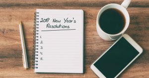 Foto de um caderno escrito resoluções de ano novo, uma xicara ao lado, um celular e uma caneta